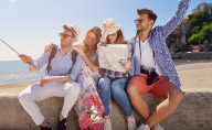 Grupa osób siedząca w słoneczny dzień na murku przy plaży.